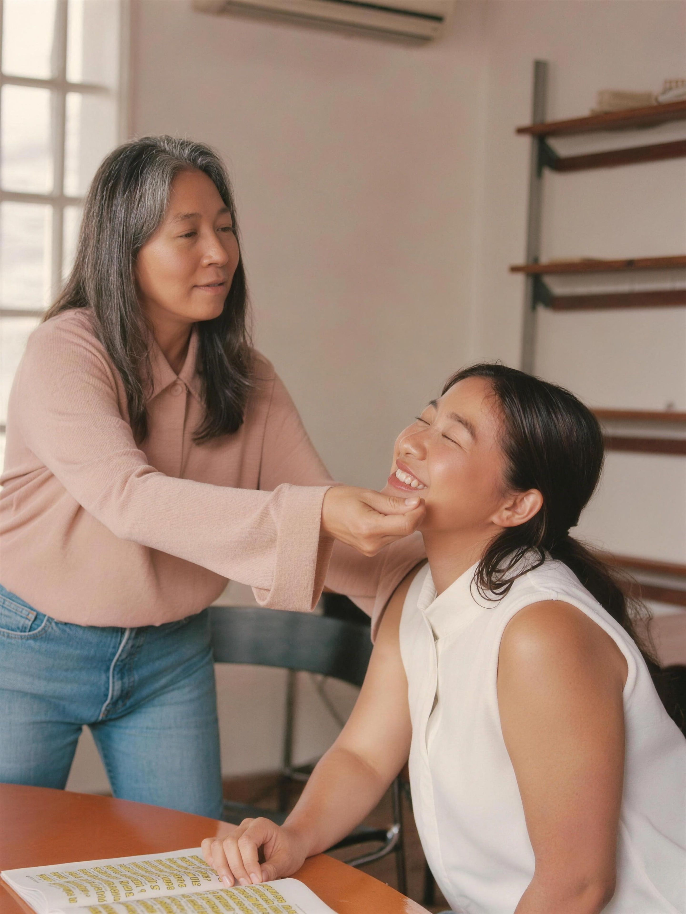 An East Asian woman mentor in her 50s with a gray bob giving warm feedback to a young Latina student across a wooden table, both smiling, bright