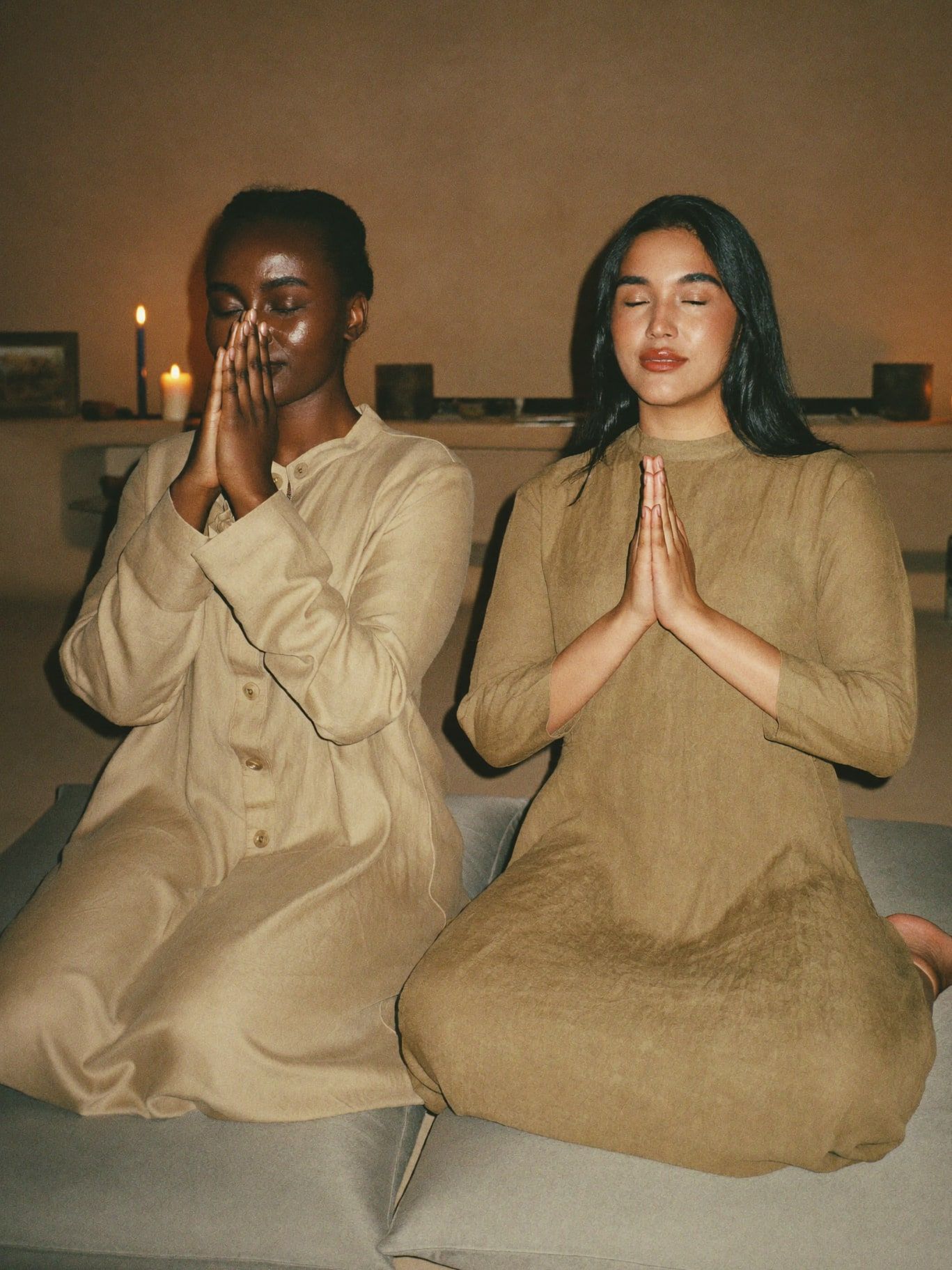 A Black woman and a South Asian woman, both wearing long-sleeved linen tunics in sand and olive, seated on floor cushions side by side chanting with
