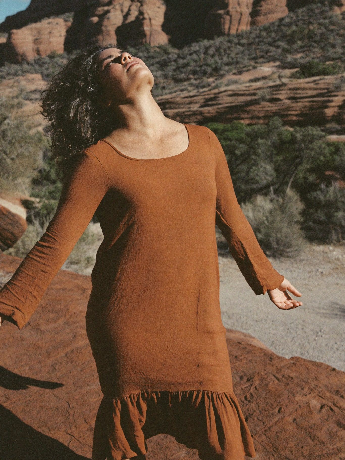 A Native Hawaiian woman in her 30s wearing a long-sleeved rust linen dress, standing alone on red rock with arms relaxed at her sides and eyes