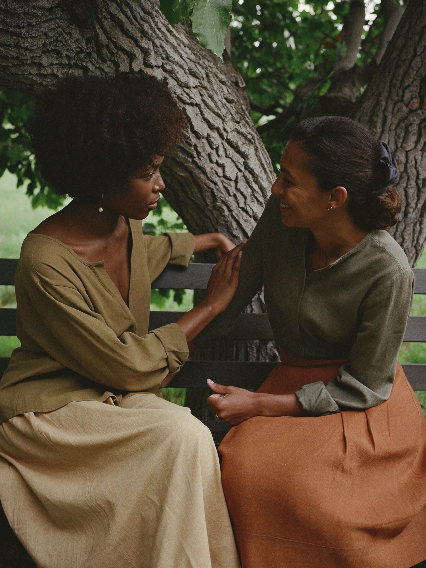 A Black woman and a Mexican American woman in a heartfelt conversation on a wooden bench under a cottonwood tree, one listening with care. Modest