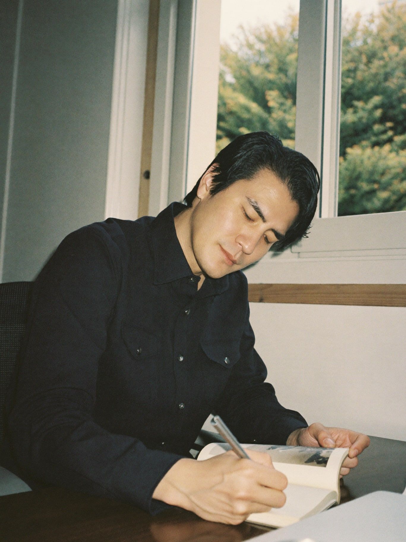 A Korean American man in his 30s writing thoughtfully in a journal at a desk by a window