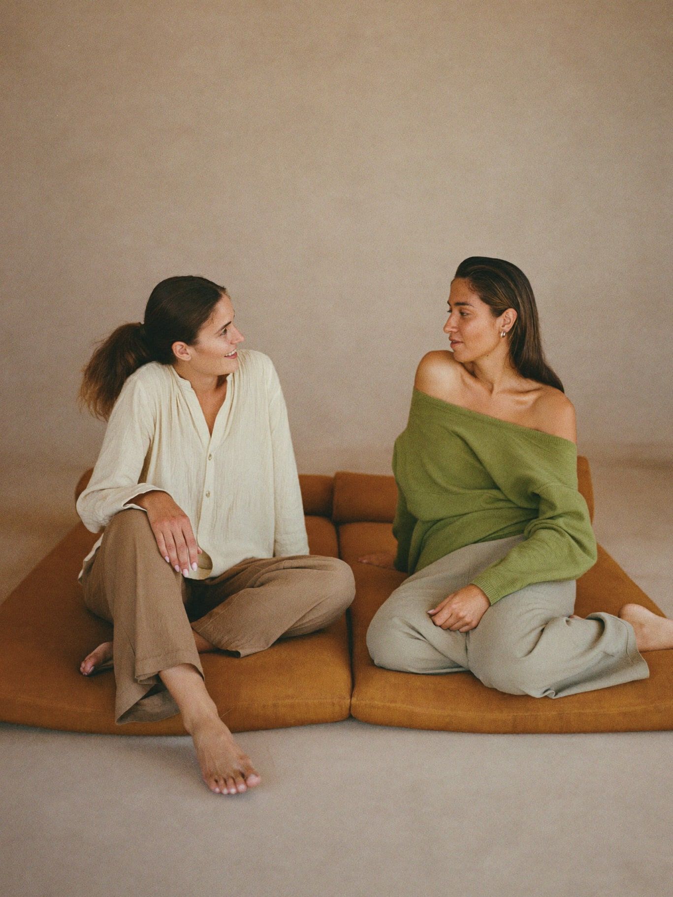 Two women of color sitting on floor cushions in a warm adobe room, one speaking softly and one listening. Modest clothing: loose long-sleeved linen