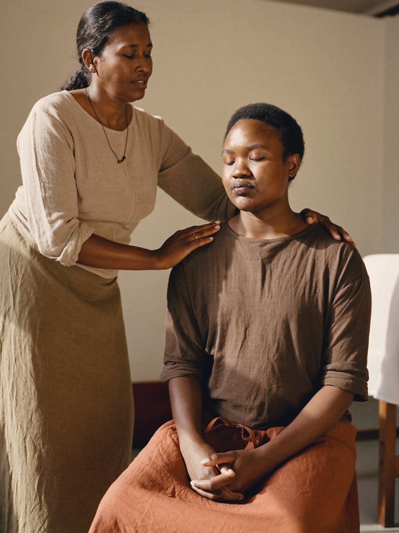 A South Asian woman healer in her 40s resting her hands lightly on the shoulders of a seated Black student with closed eyes, practicing energy