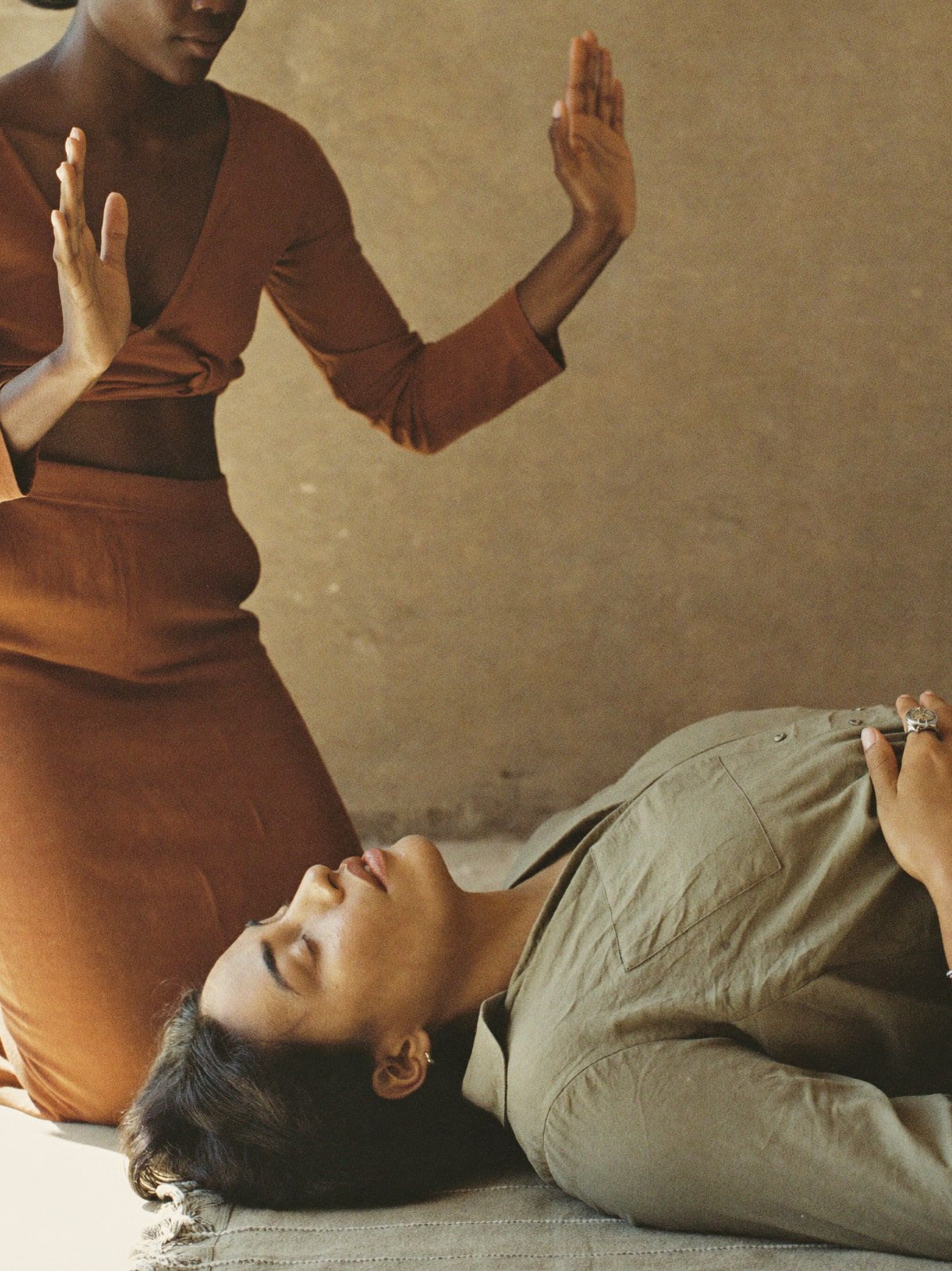 A Latina woman lying on a linen mat with eyes closed while a Black woman kneels beside her, palms hovering above her, practicing energy healing in a