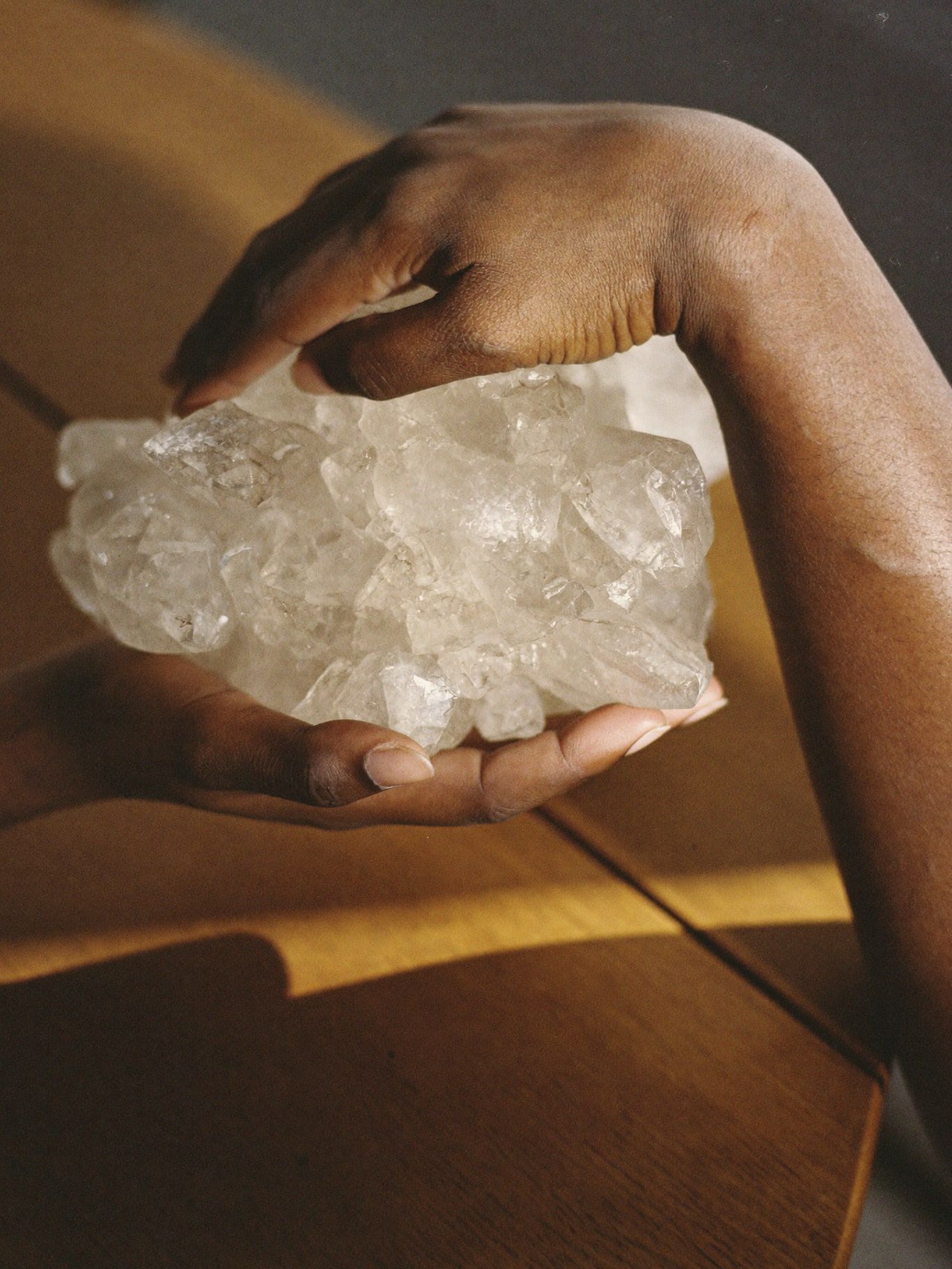 Brown hands hovering above a large clear quartz cluster on a wooden table, warm window light