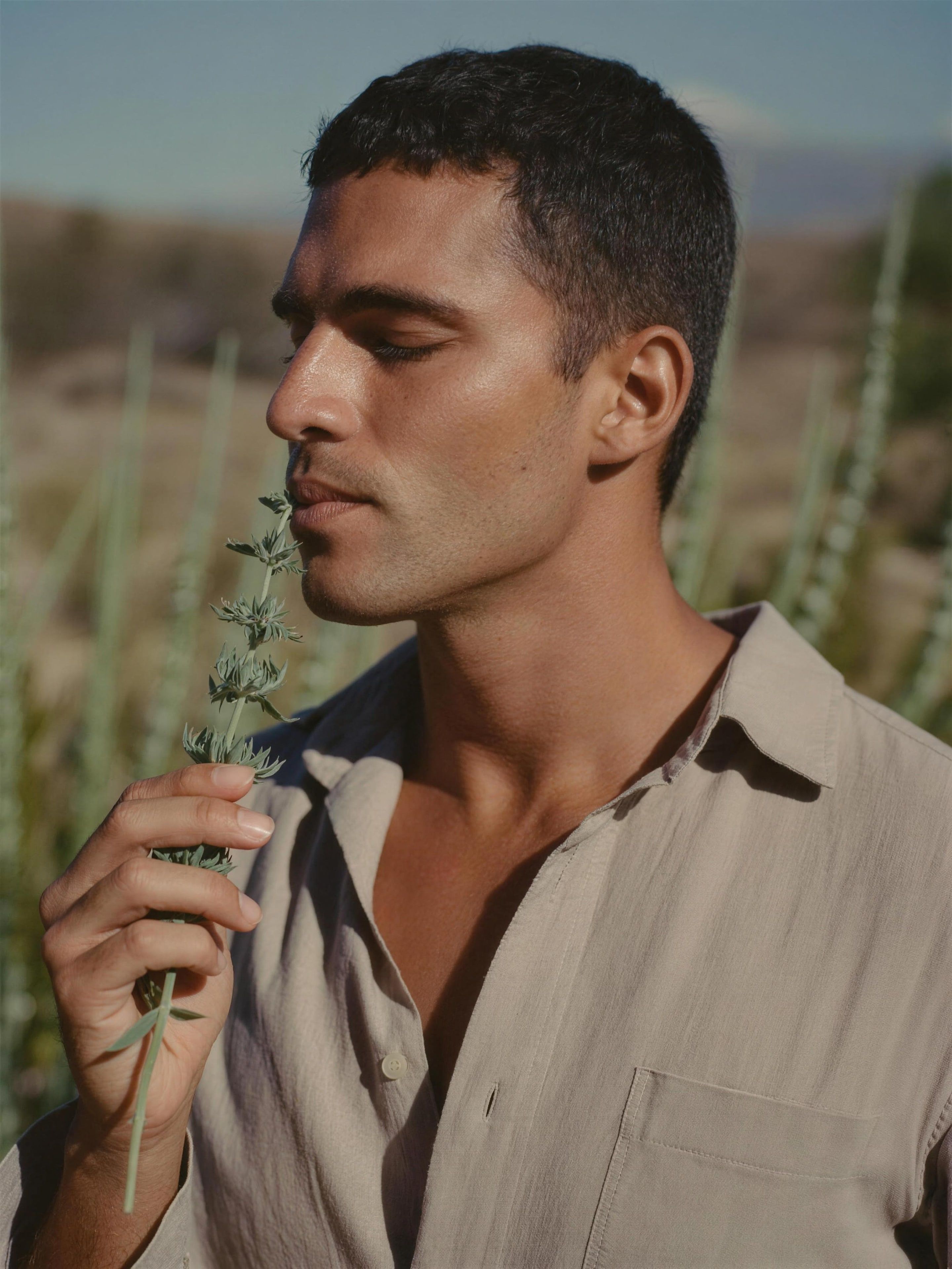 A Latino man in his 30s wearing a buttoned sand linen shirt, eyes closed, holding a sprig of sage near his face and breathing in its scent, desert