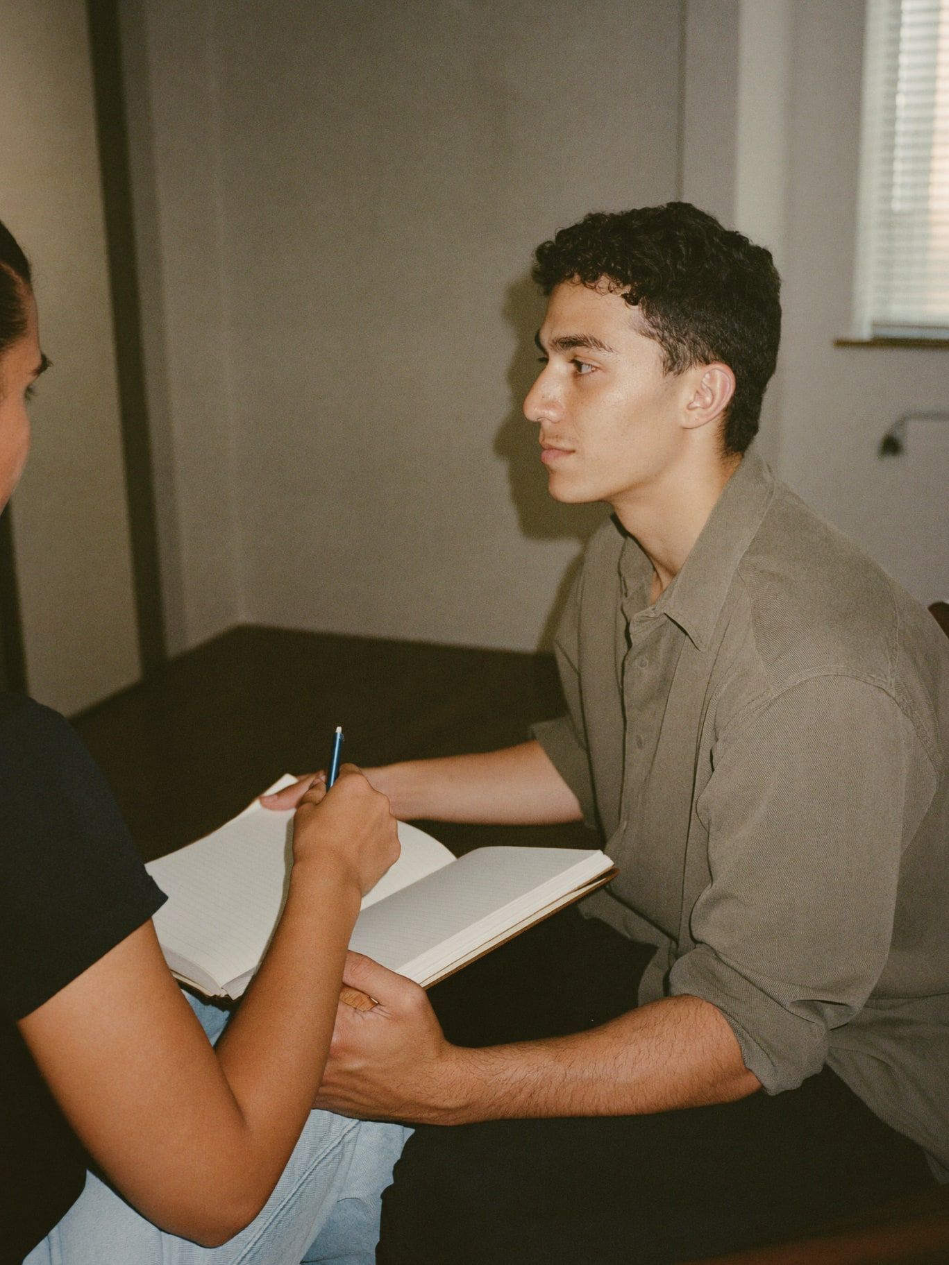 A young Latino man in an olive shirt, a trainee energy reader, sitting with a client in a calm reading room, a notebook on his knee, listening