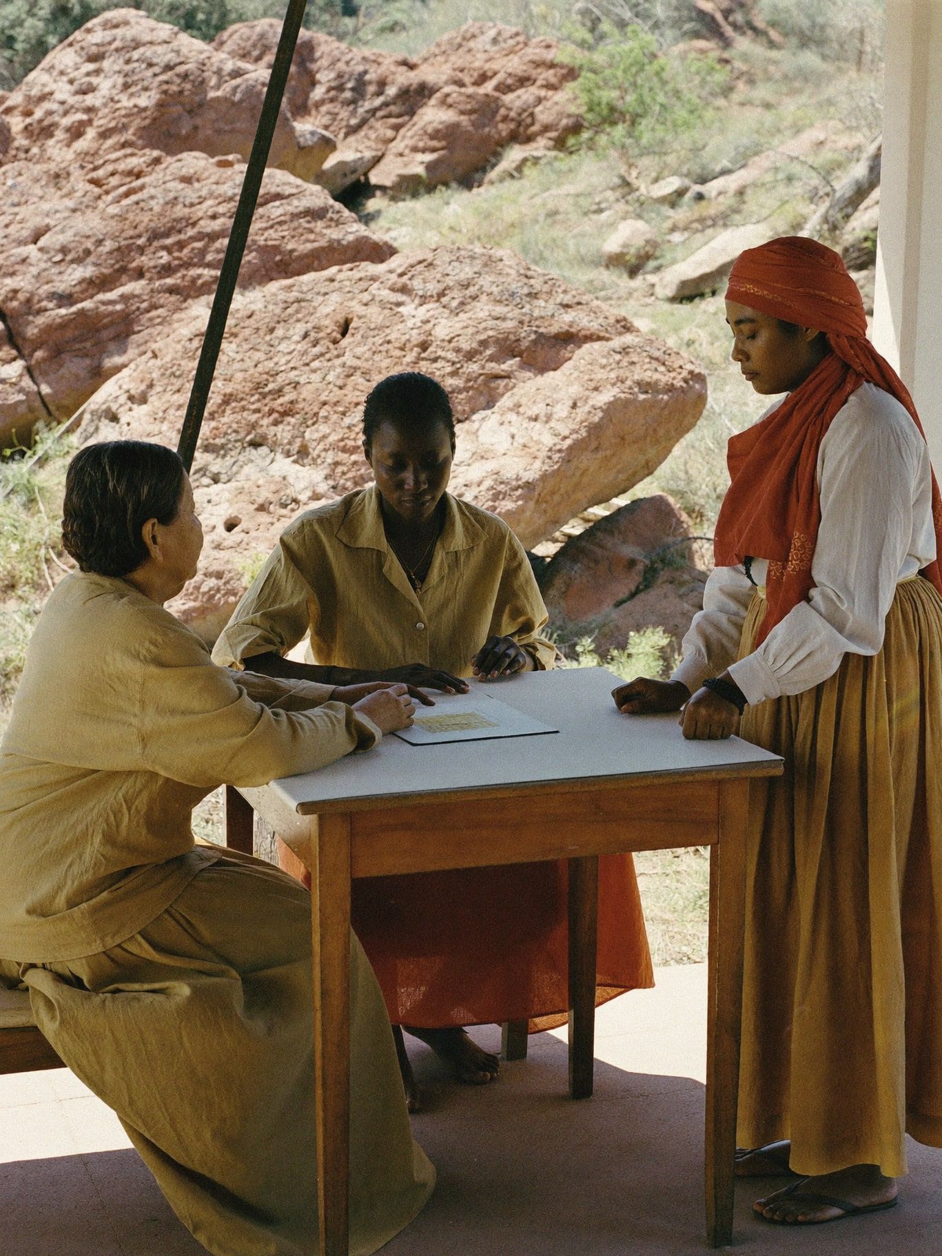 A Black woman and a Filipina woman practicing an intuitive reading at a small table on a shaded patio, red rocks behind them, afternoon light. Modest