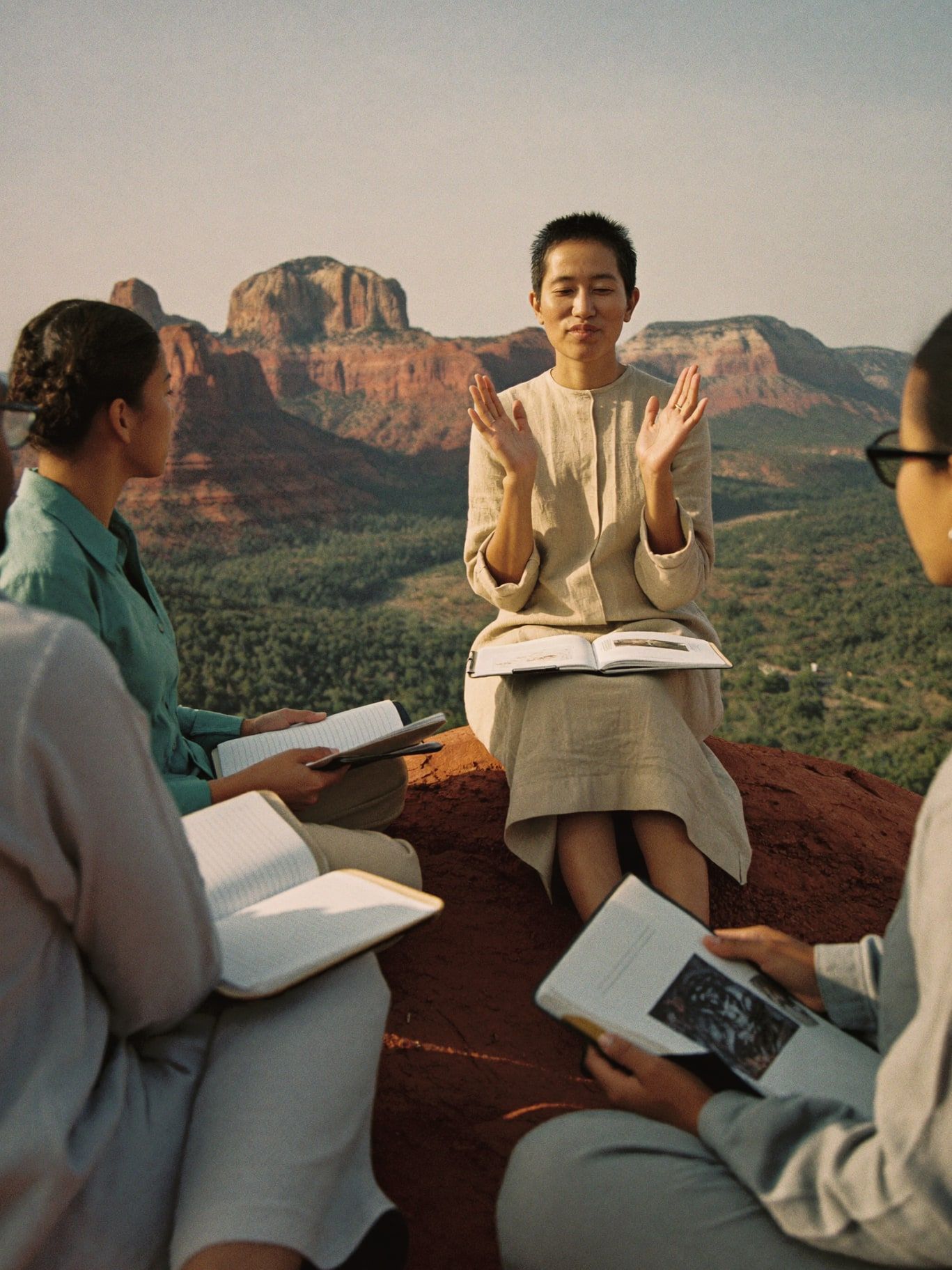 A trainer in a sand linen tunic demonstrating an energy reading to trainees seated in a circle on red rock, buttes behind them