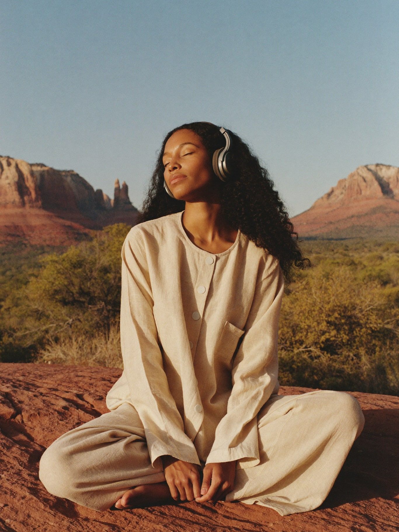 A woman in a long-sleeved linen tunic listening with headphones, eyes closed, seated on red rock at dawn