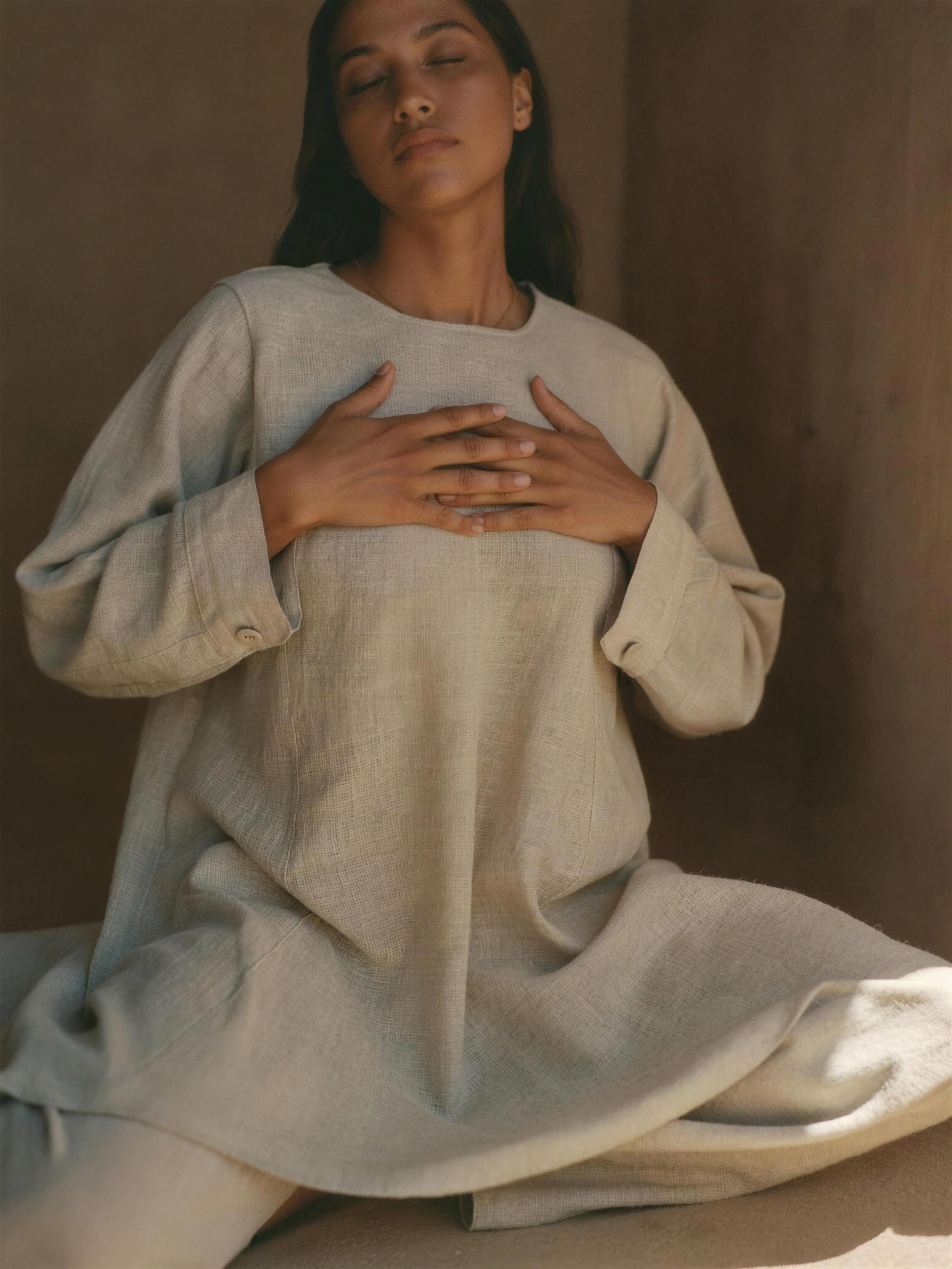 A Latina woman in her 30s wearing a long-sleeved sand linen tunic and wide trousers, sitting cross-legged in a sunlit adobe room with both hands