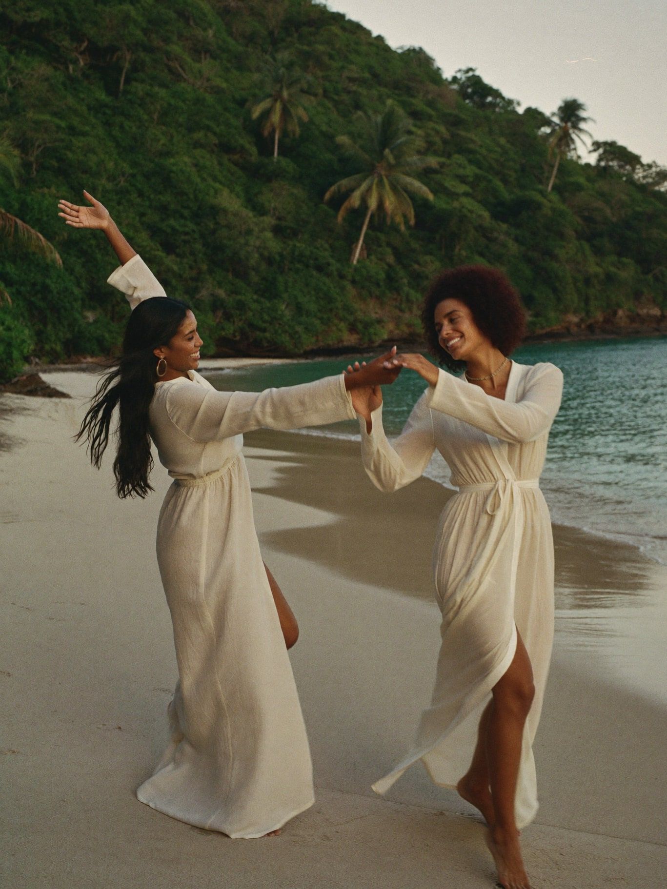 Two women, one Black and one Latina, wearing long flowing long-sleeved linen dresses, dancing barefoot on a jungle-edge beach in Costa Rica at
