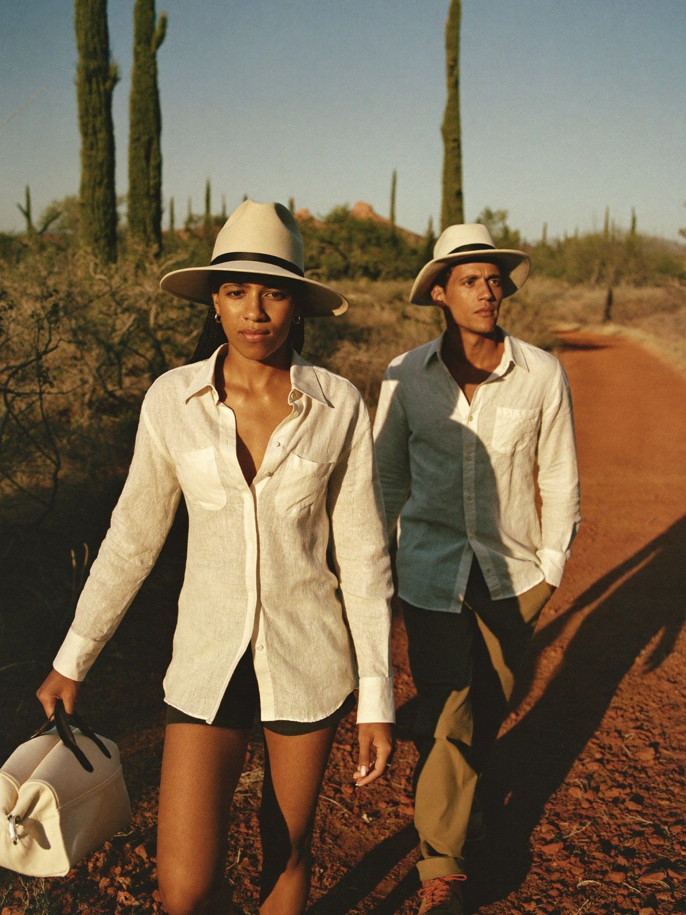 A Black woman and a Latino man, both wearing long-sleeved linen shirts, wide-brim hats and hiking trousers, walking a red dirt trail through junipers