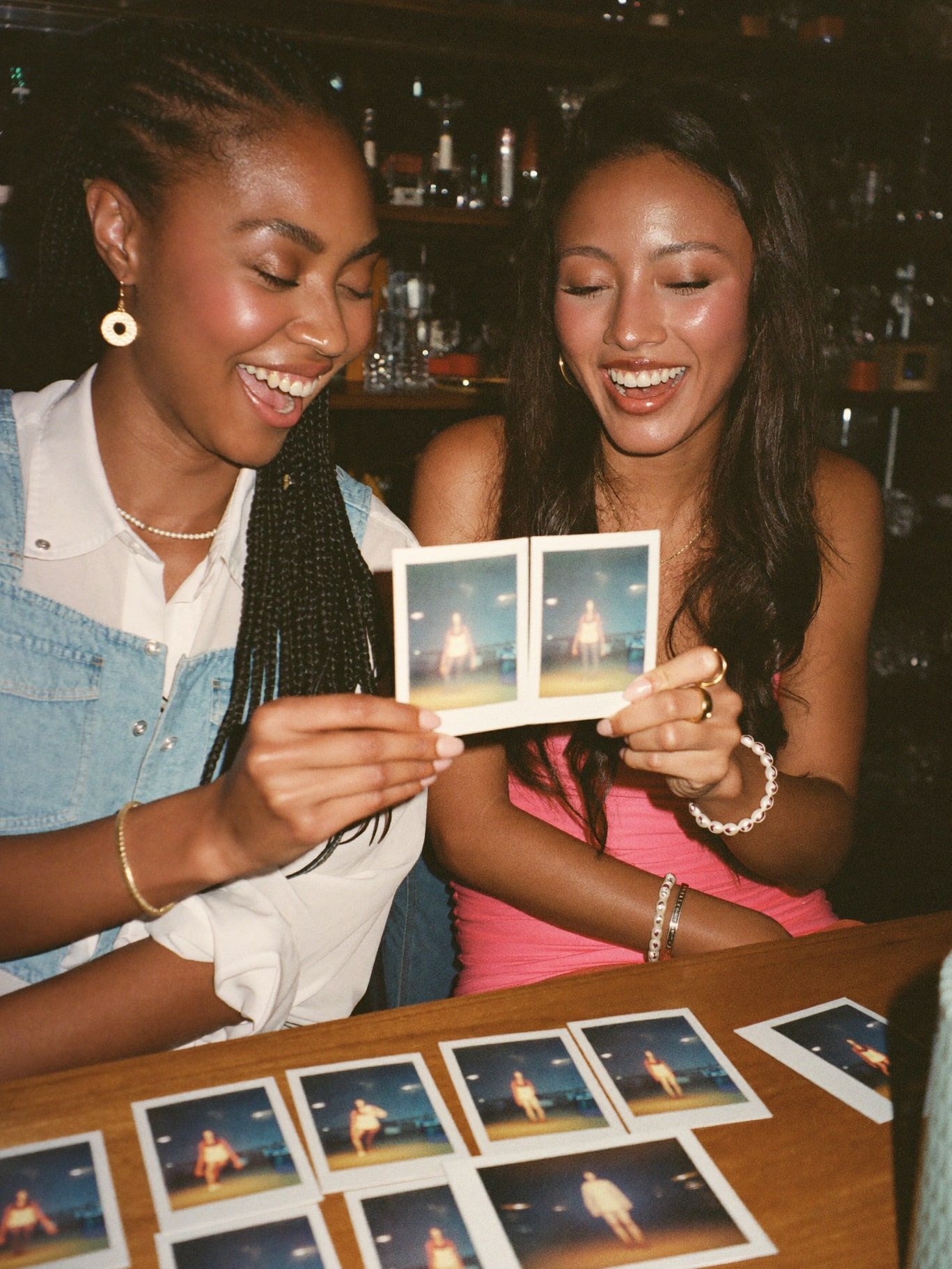 Two young women, one Black and one Mexican American, laughing together as they compare their glowing instant aura photographs at a wooden table in a