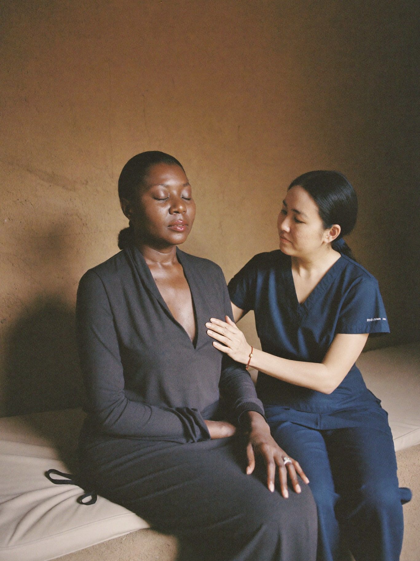 A Black woman in her 40s sitting on a cushioned bench with eyes closed and one hand over her heart, a Filipina practitioner sitting beside her with