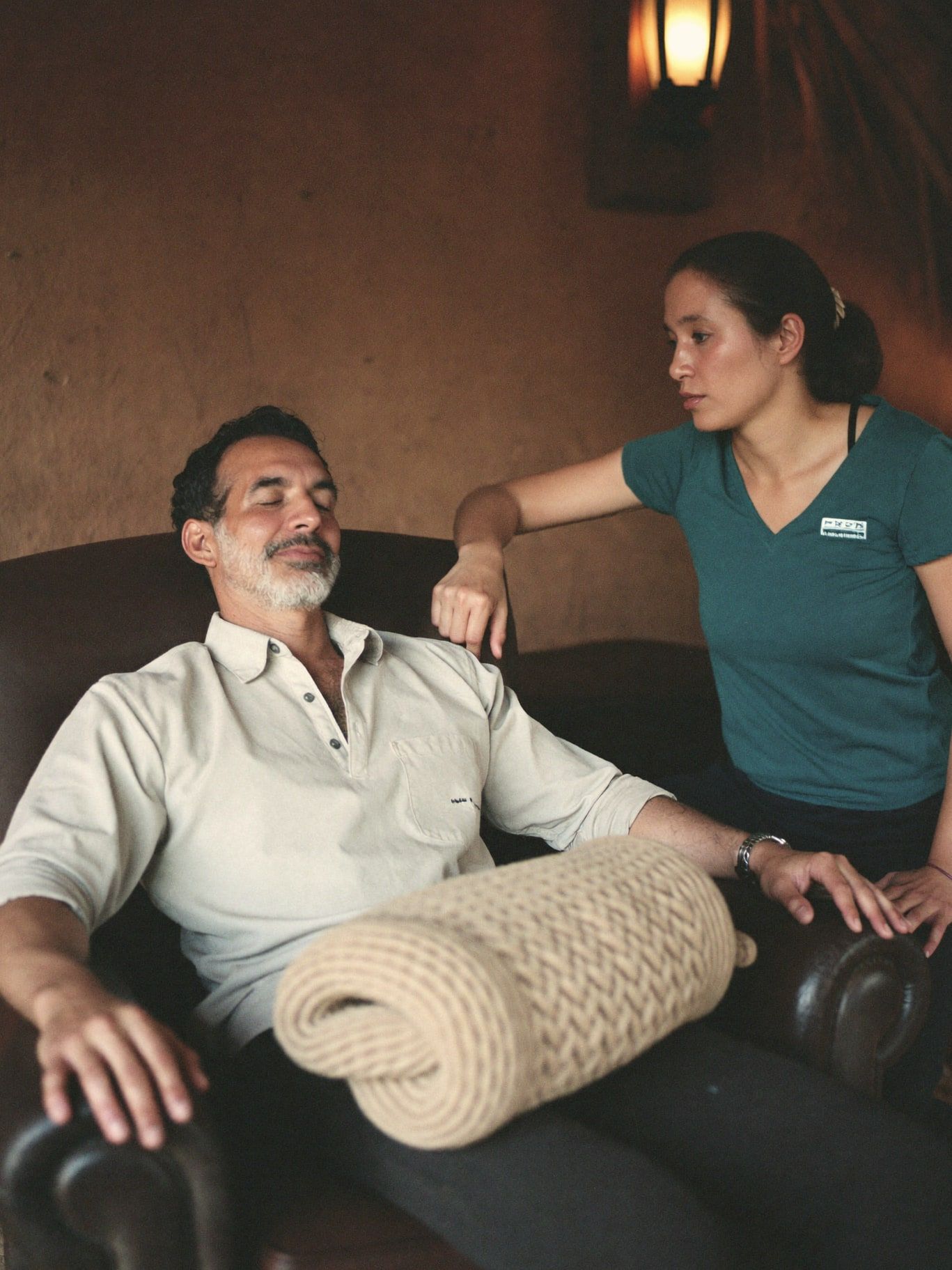 A South Asian man in his 50s reclining in a deep armchair with eyes closed and a wool blanket over his knees, a Latina guide in her 40s seated beside