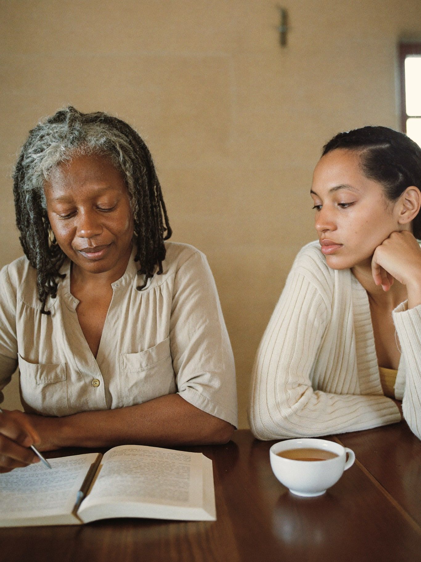 A Black woman intuitive reader in her 50s with silver-streaked locs, wearing a sand linen shirt, sitting across a small wooden table from a young