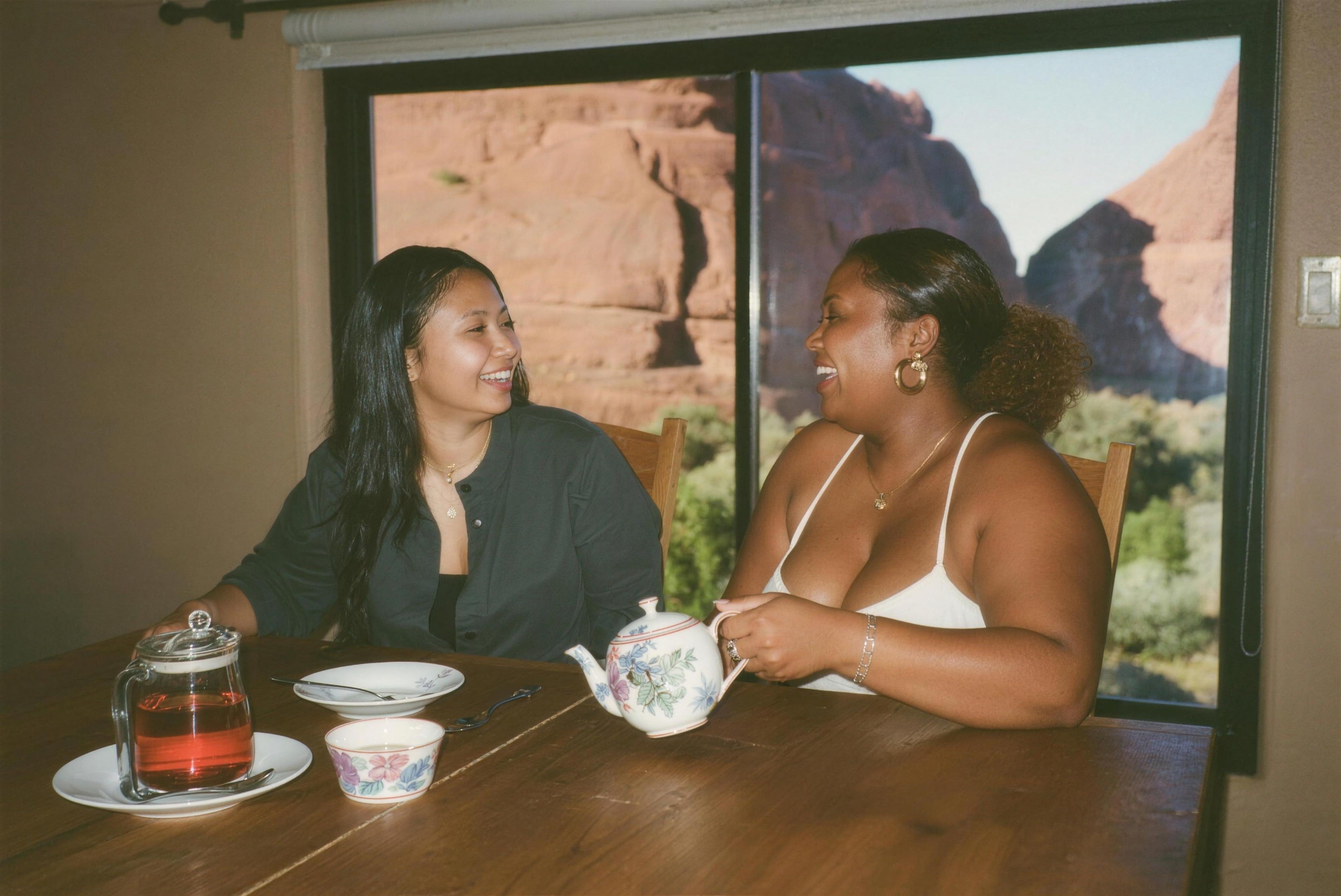 Two friends laughing over tea, red rocks through the window