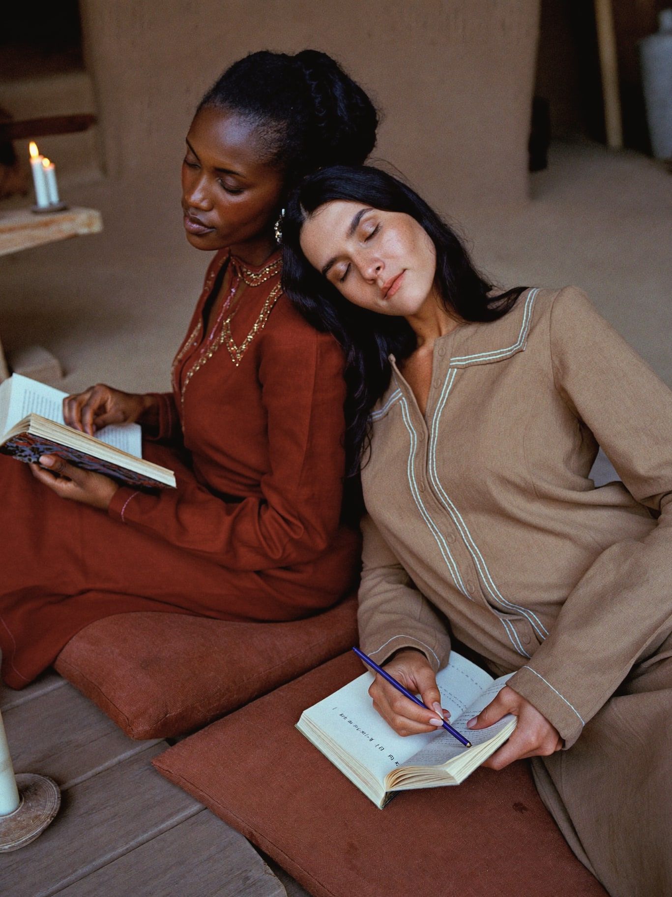 A Black woman and a Latina woman, both wearing long-sleeved rust and sand linen tunics, seated on cushions by candlelight with notebooks open on